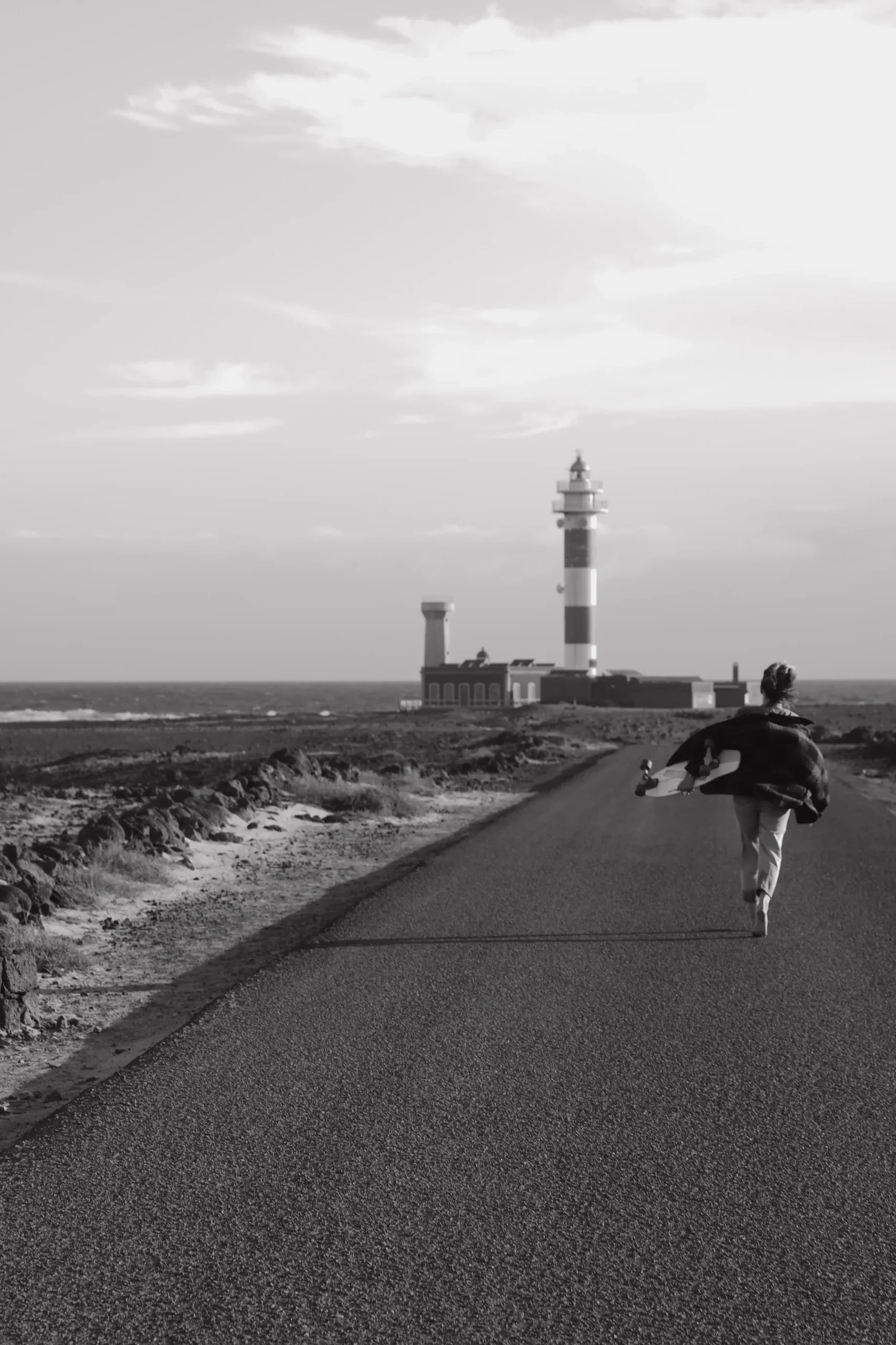 Lina läuft mit einem Skateboard auf einer Küstenstraße in Richtung Leuchtturm