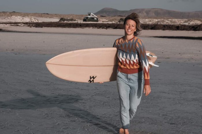Lina Leppke mit Surfboard unter dem Arm, läuft durch das Wasser, Blick in die Kamera, im Hintergrund sieht man einen Campervan und die Berge Fuerteventuras